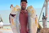 Brad Sutcliffe holds two large fish up to the camera, he is out on the water with the horizon in the background.