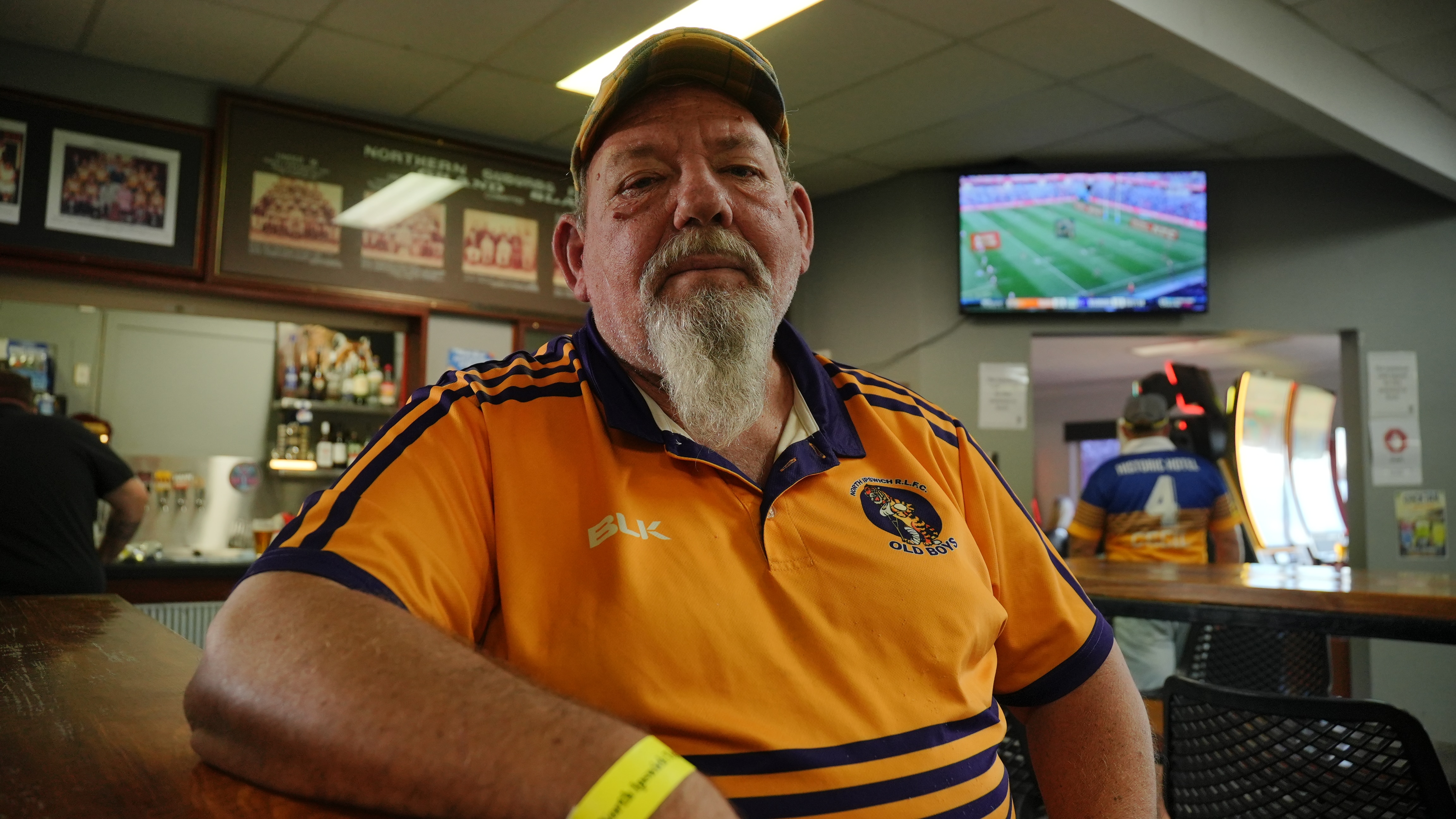 Low-angle shot of a man with a long white-goatee and a yellow footy polo top. He is sitting inside a club-house bar.