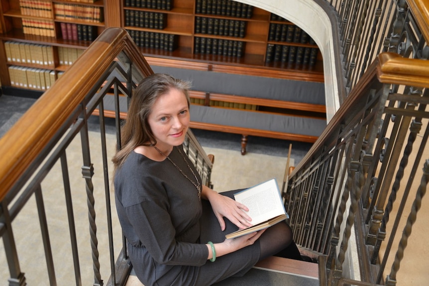 A woman holding a book sits in a staircase in a library.
