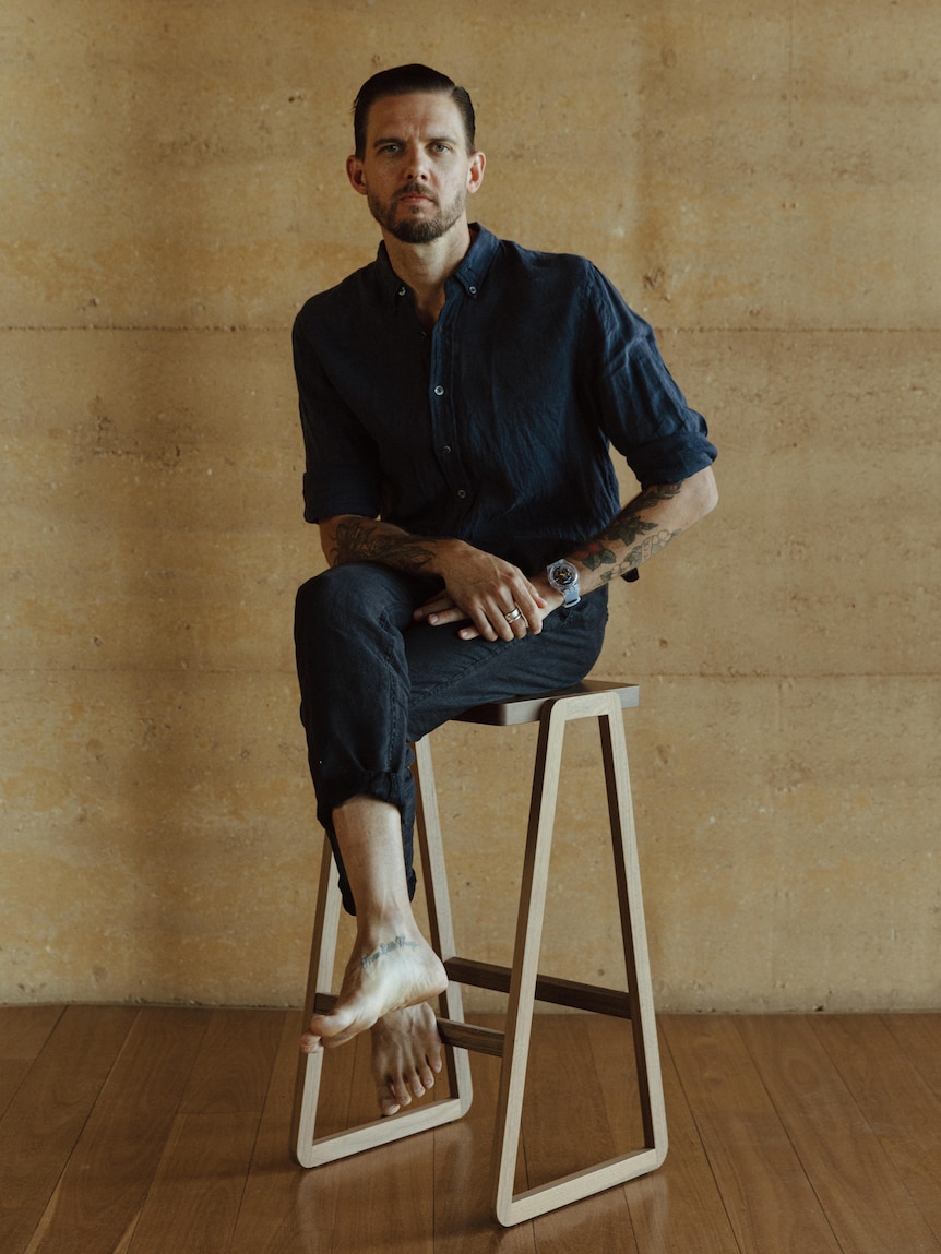 A man with short dark hair sits on a stool with his legs crossed, looking thoughtful.