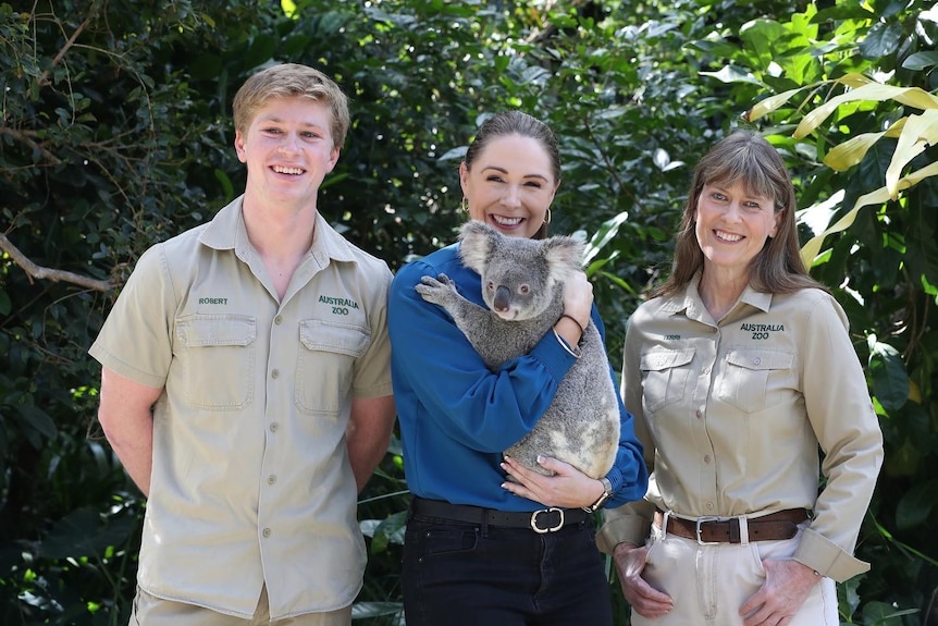 Three adults stand smiling with a koala