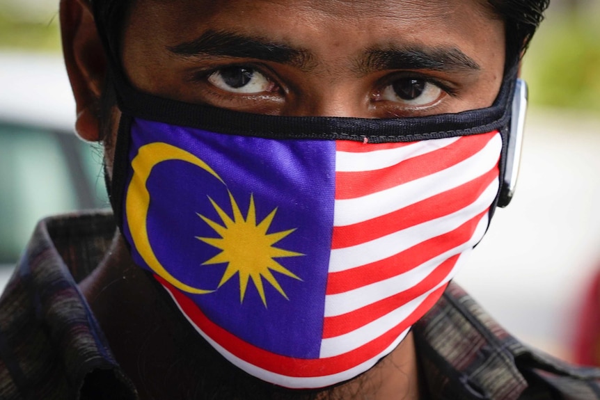 A close up of a man with brown eyes staring at the camera wearing a face mask with the Malaysian flag printed on it.