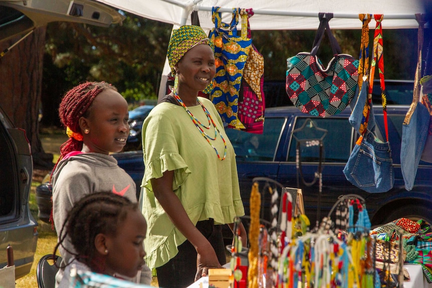a woman and two girls stand at a market stall surrounded by colourful bags