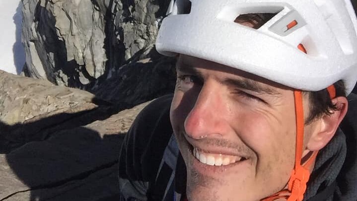 a photo of a man smiling at the camera with a helmet on and a rockface in the background