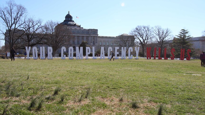 Giant red and white letters spelling out "Your Complacency Kills Us" stand across from the US capitol