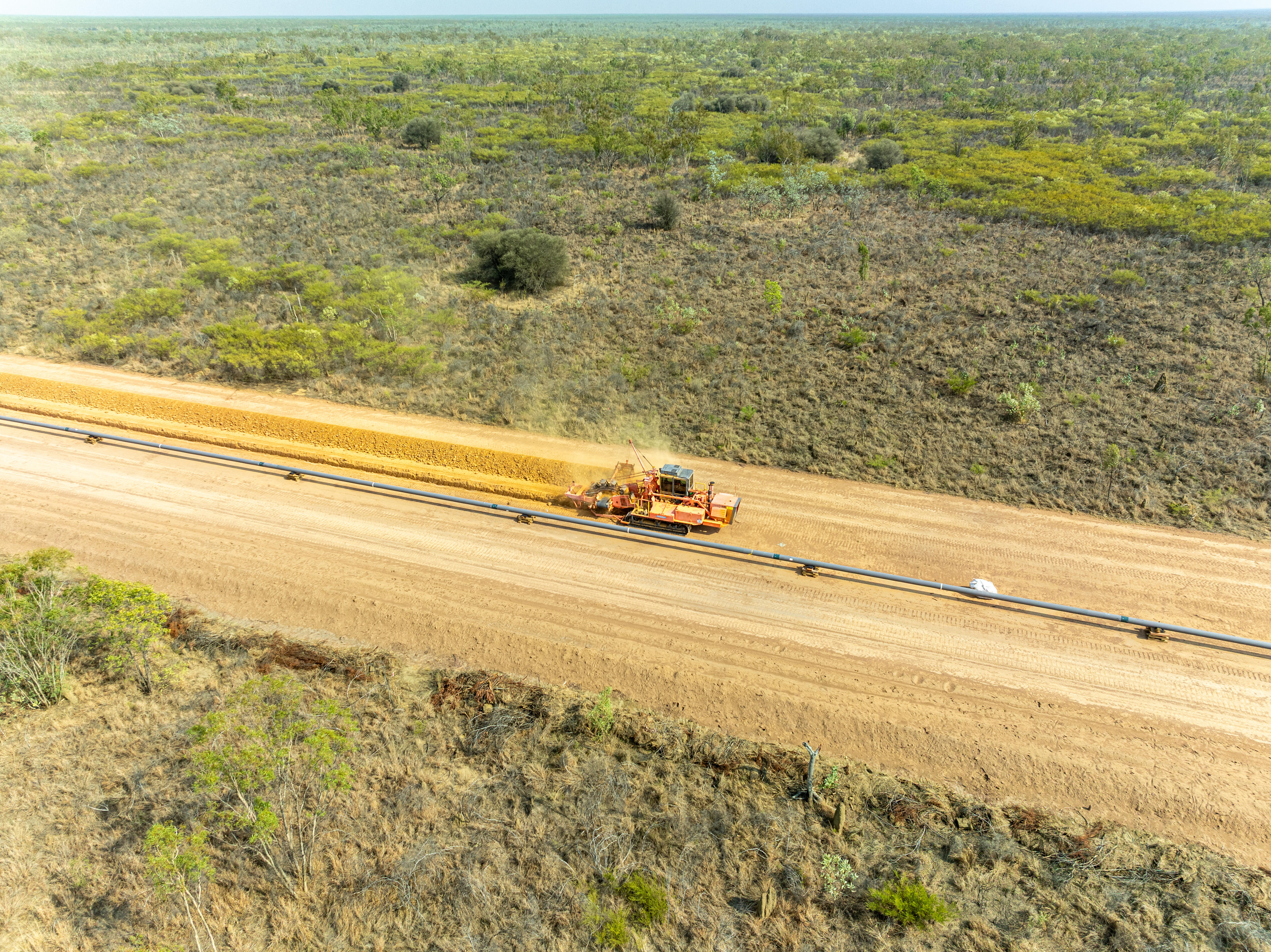 Aerial shot of yellow grader machine moving across dirt surface, with gray pipeline visible from left to right