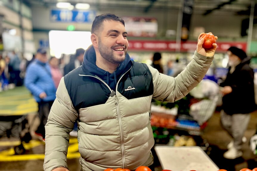 a man in a puffer jacket holds up an apple in a busy produce market
