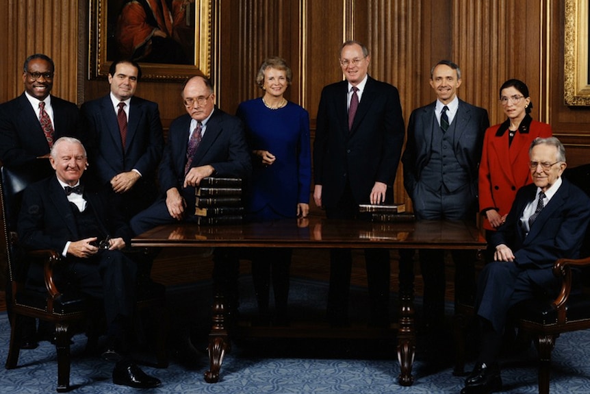 Colour photo of the 1993 Supreme Court Justices posing in front of a wooden table and and room with ornately framed paintings.