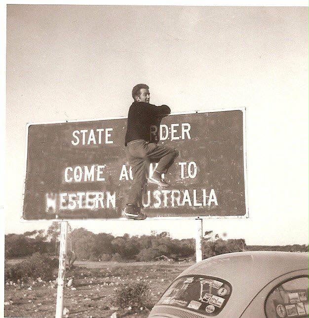 Black and white photo of Lance Sturtzel hanging on the West Australian state border sign.