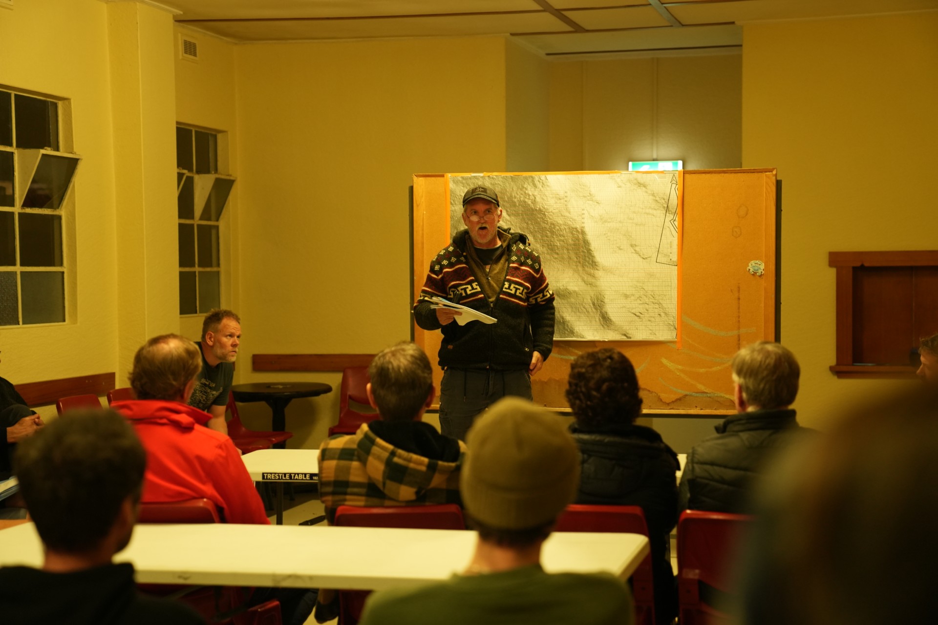A man with grey hair and glasses standing while people sit and listen