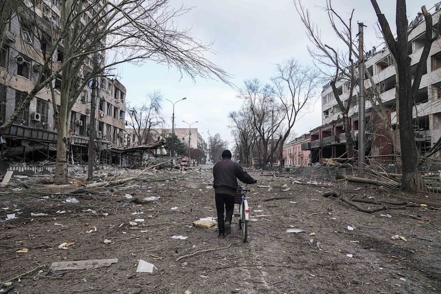 An old man pushes a bike down a street with damaged buildings on each side