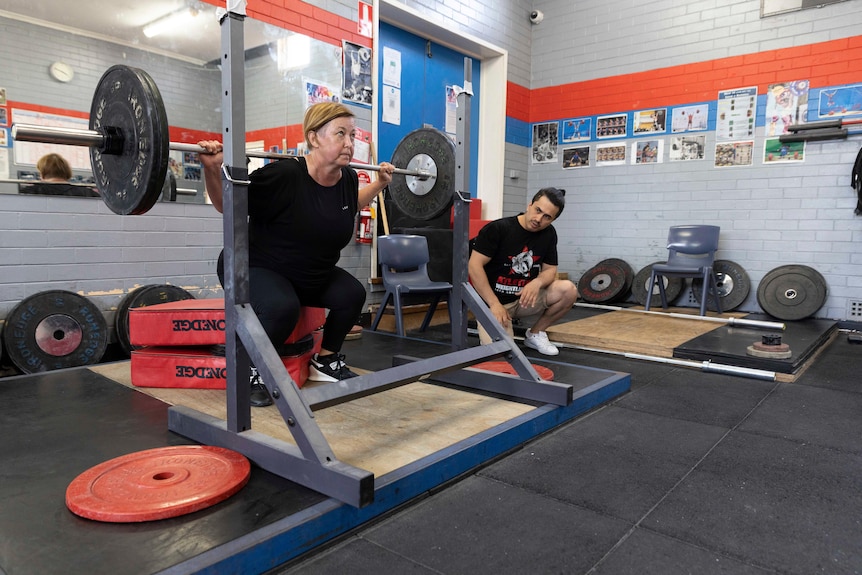 A middle-aged woman lifts weights while a man, her coach, watches on.