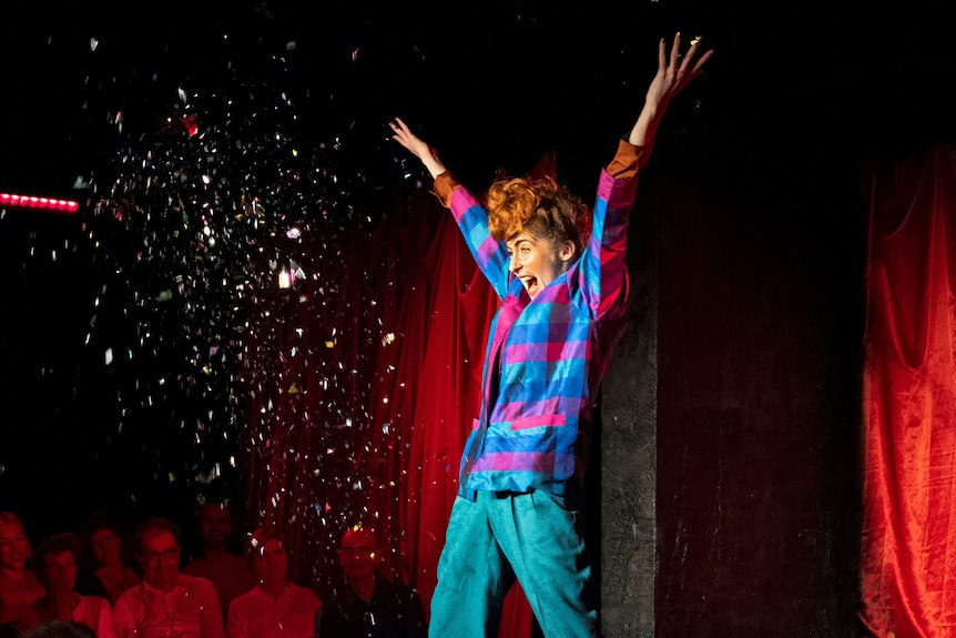A woman in her late 30s in colourful clothes on stage, her arms are raised and glitter is being shot at her