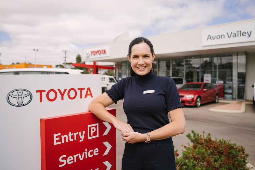 A woman in a navy top and skirt smiling, leaning against a Toyota sign in front of a row of shops on a cloudy day.