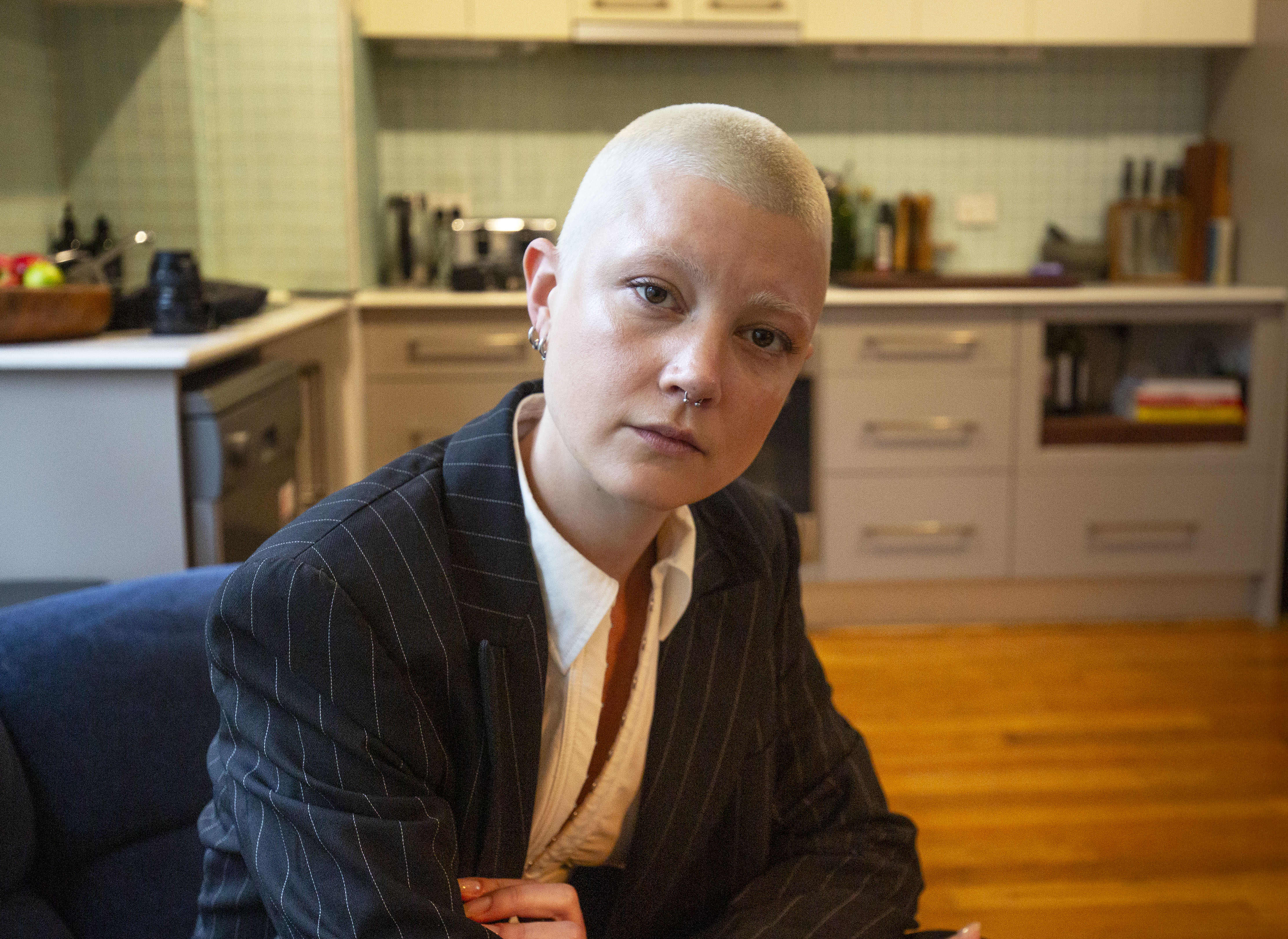 A woman with short blonde hair, wearing a pinstripe jacket, sits in the kitchen of a house.
