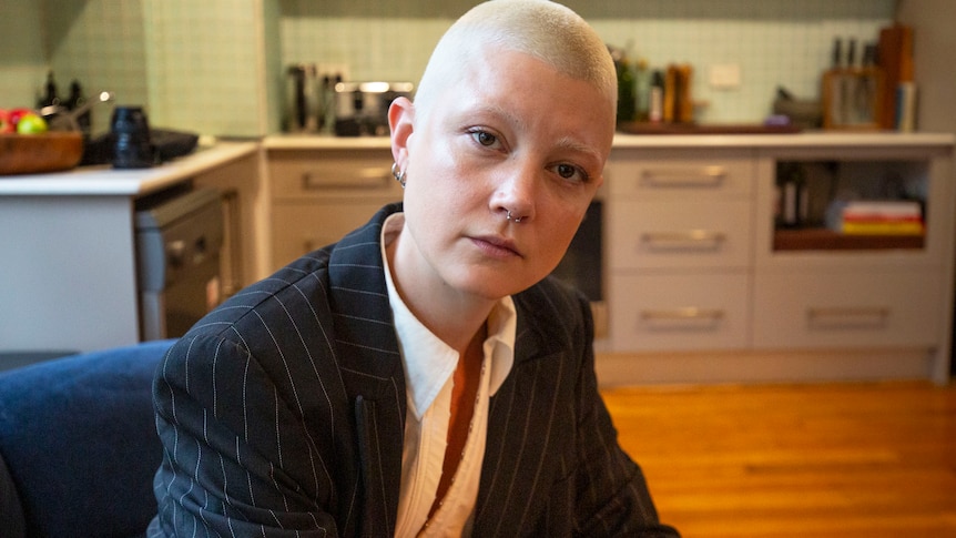 A woman with short blonde hair, wearing a pinstripe jacket, sits in the kitchen of a house.