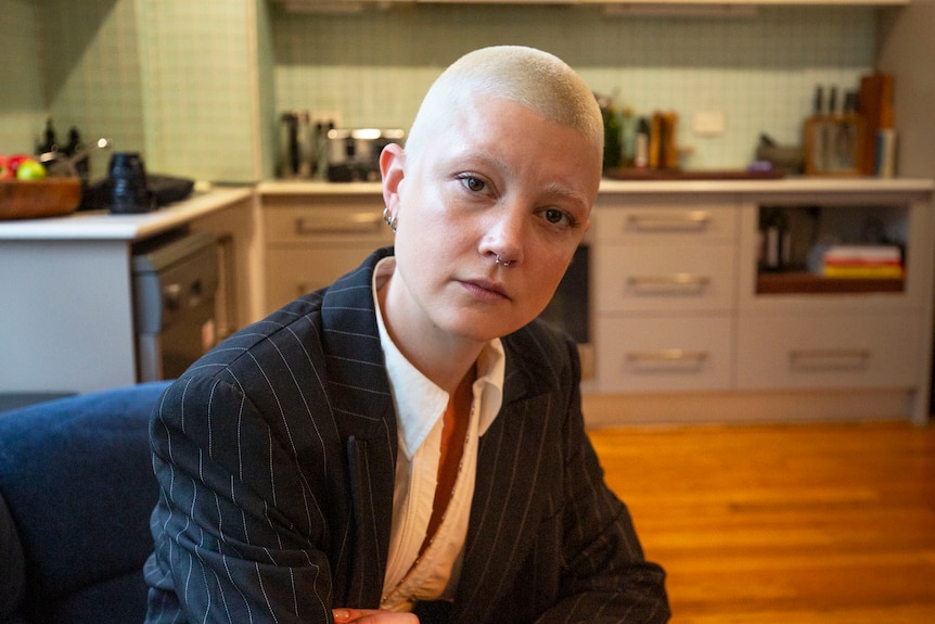 A woman with short blonde hair, wearing a pinstripe jacket, sits in the kitchen of a house.