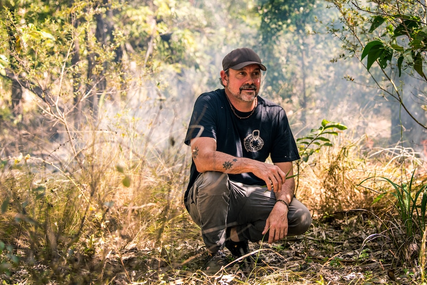 A man crouching down in Australian bush while light smoke from a small fire rises behind him