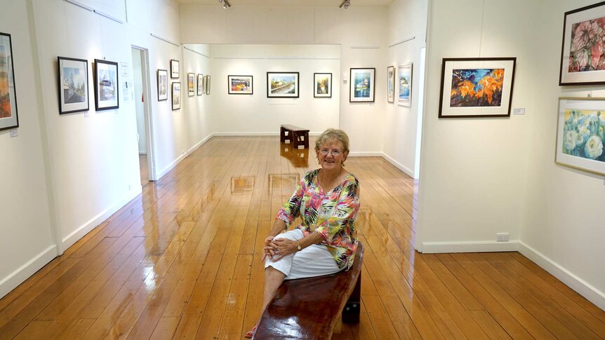 Elaine Madill sits in the middle of an art gallery space surrounded by paintings