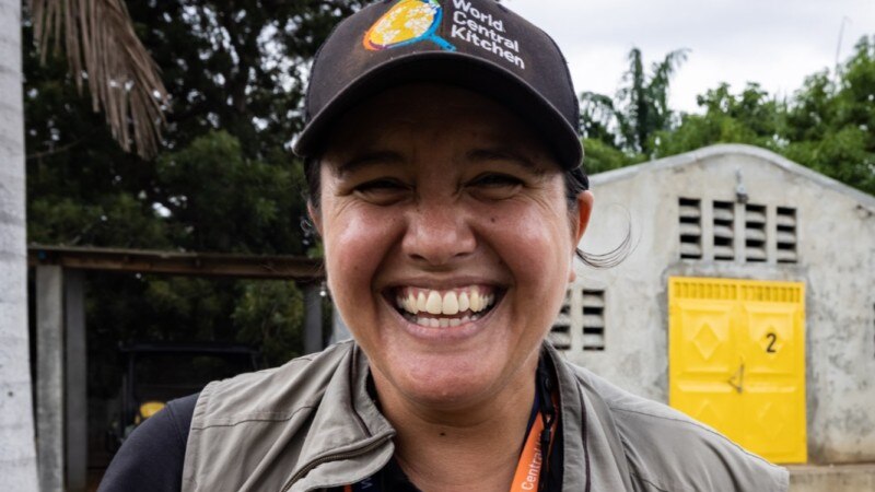 A woman wearing a black cap and a khaki vest smiles at the camera
