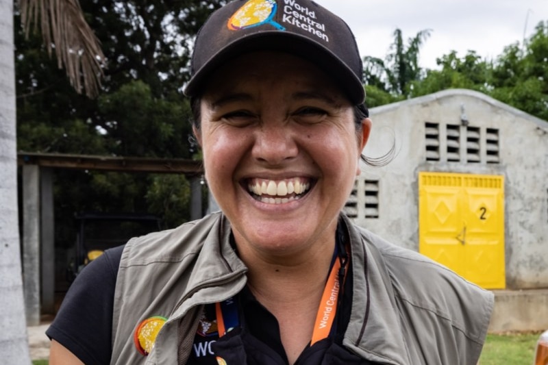 A woman wearing a black cap and a khaki vest smiles at the camera