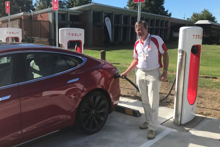 A man stands next to his Tesla electric vehicle, charging it with electricity in a carpark.