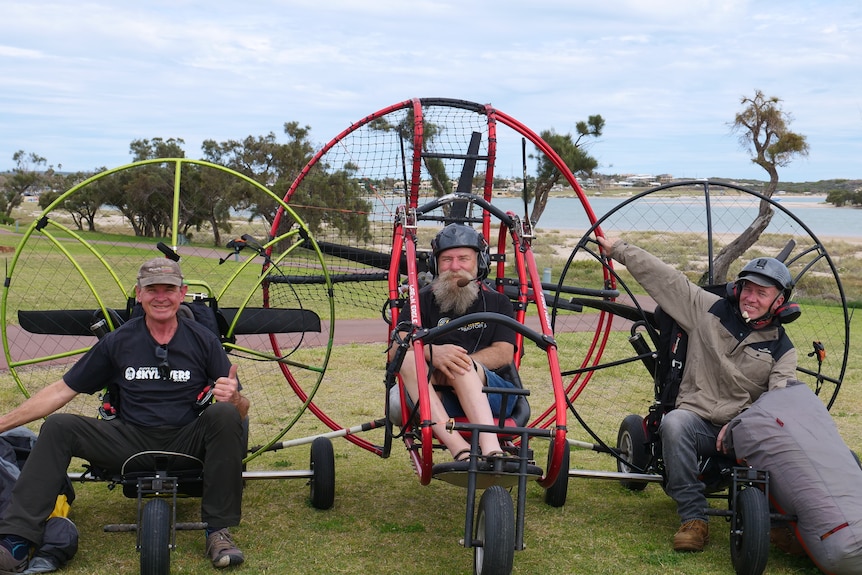 Three men smile as they sit in paramotors on the ground.