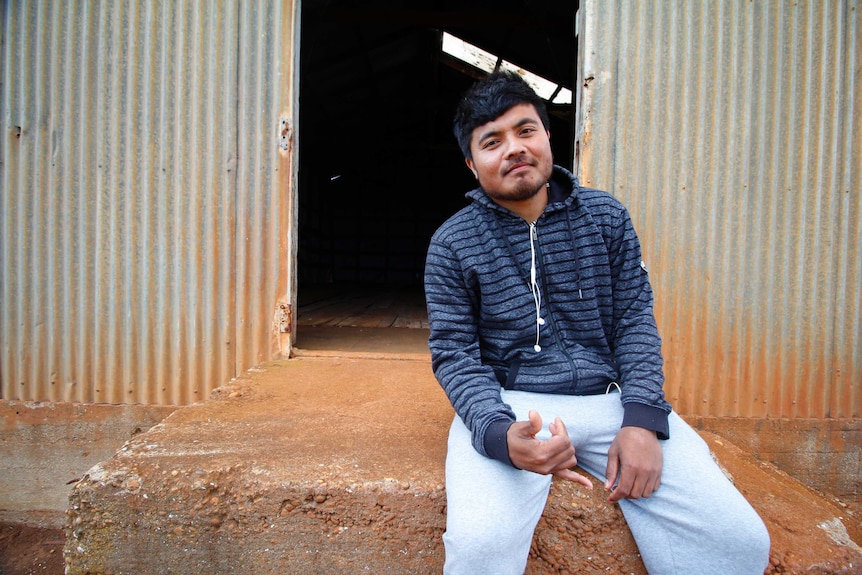 A young man smiles as he sits outside a corrugated-iron shed.