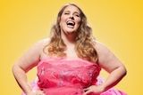 Plus-sized woman with brown wavy hair wears bright pink strapless dress, against yellow background, head thrown back, laughing.