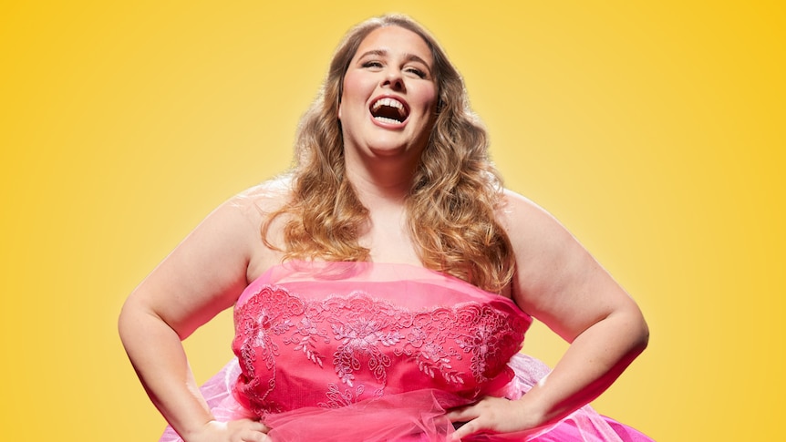 Plus-sized woman with brown wavy hair wears bright pink strapless dress, against yellow background, head thrown back, laughing.