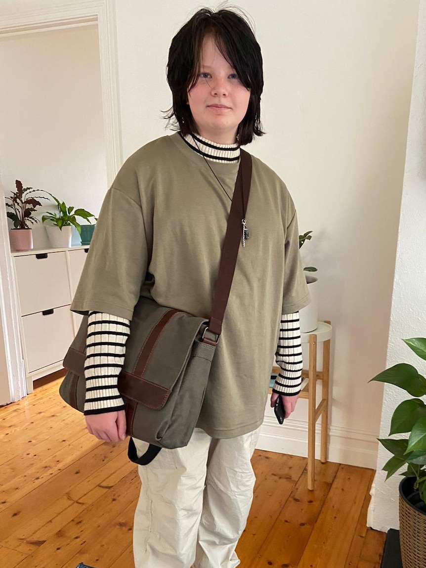 Sam in his living room, wearing an oversized shirt and a satchel bag.