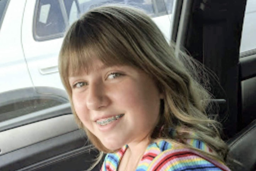 A girl with blonde hair and blue eyes wears a rainbow shirt as she smiles in a car.