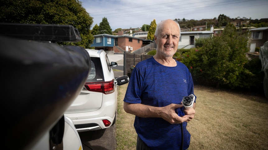 A middle aged man wearing a blue tshirt holds an electric car charger in a suburban driveway