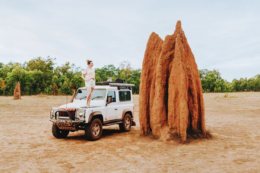A woman stands on the bonnet of a small four-wheel drive looking at a red ant hill that towers over her