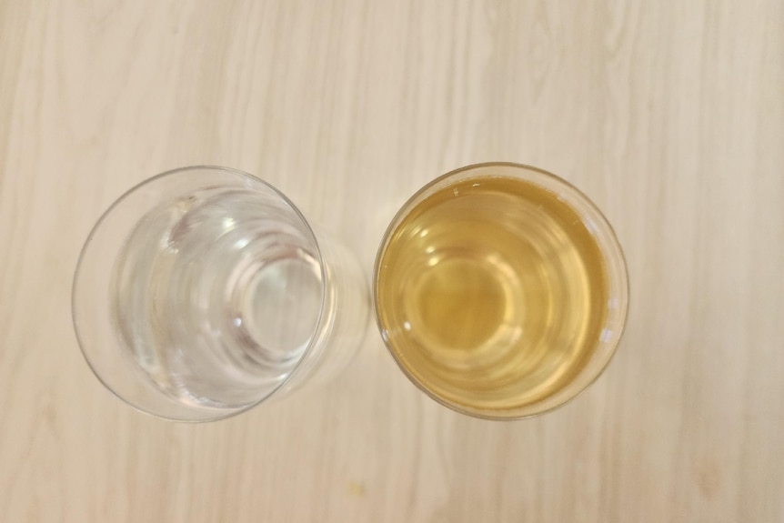 Two glasses sit side by side on a wooden table the one on the left has clear water on the right is brown. 