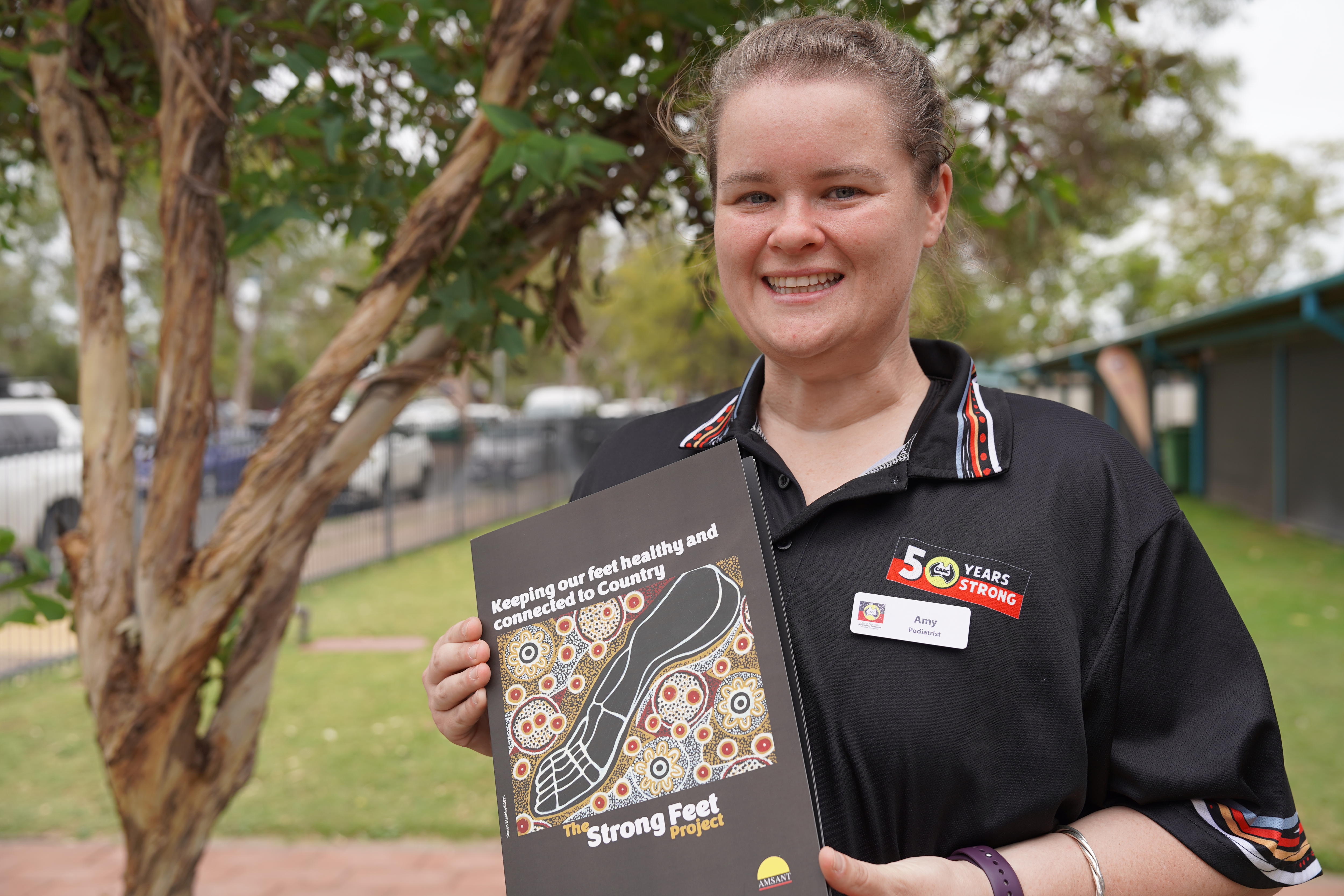 Uma mulher sorri, vestida com uma camisa pólo preta, segurando um folheto, em pé no gramado da frente de um prédio.