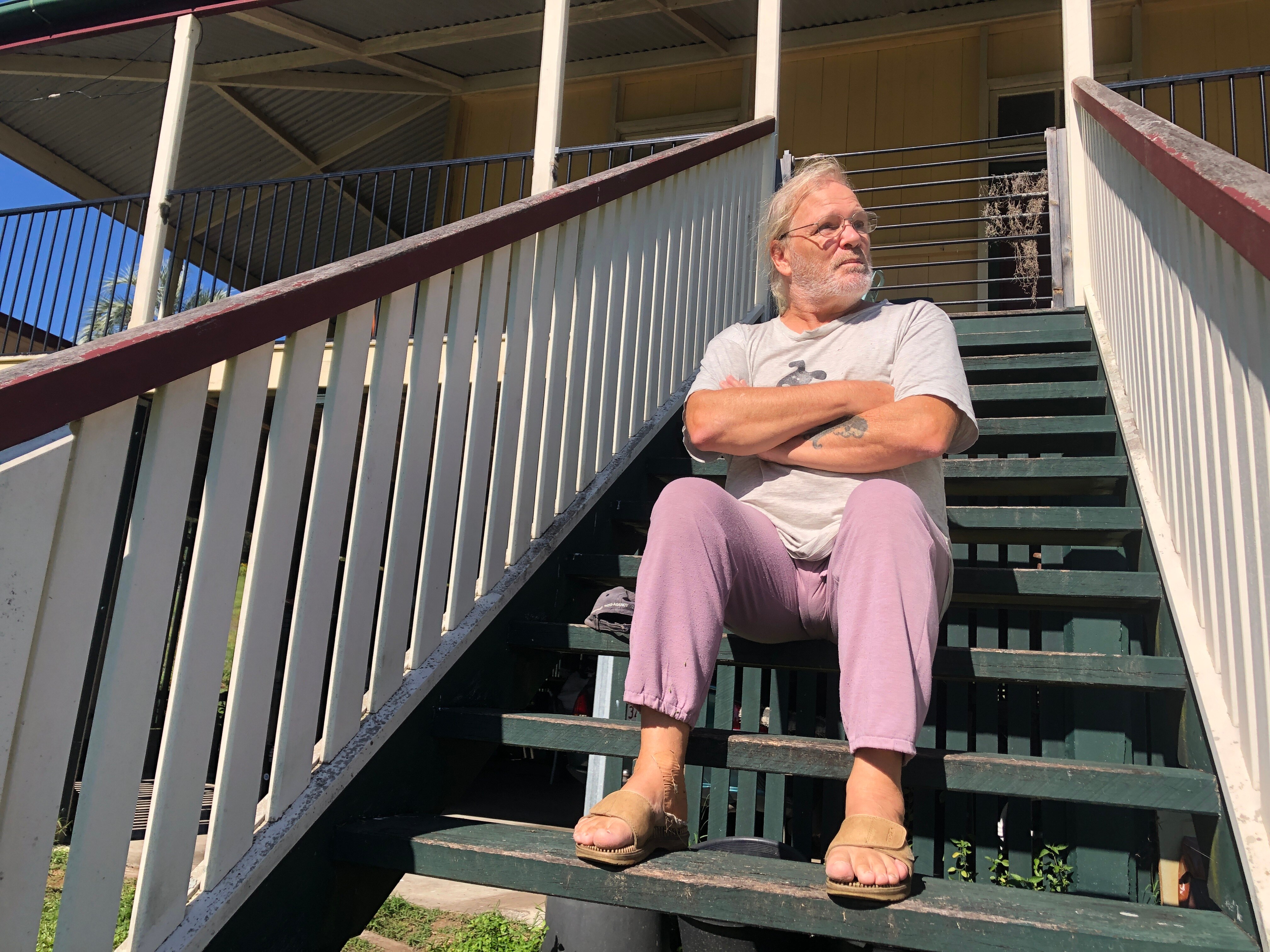 An older man sitting on the steps of an old house, his arms crossed.