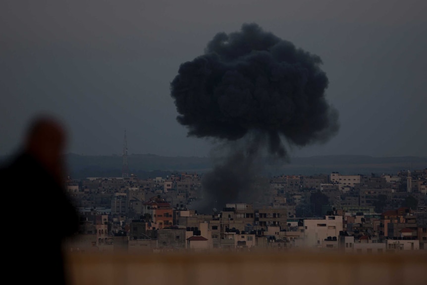 A large cloud of black smoke rises above buildings in a dark sky