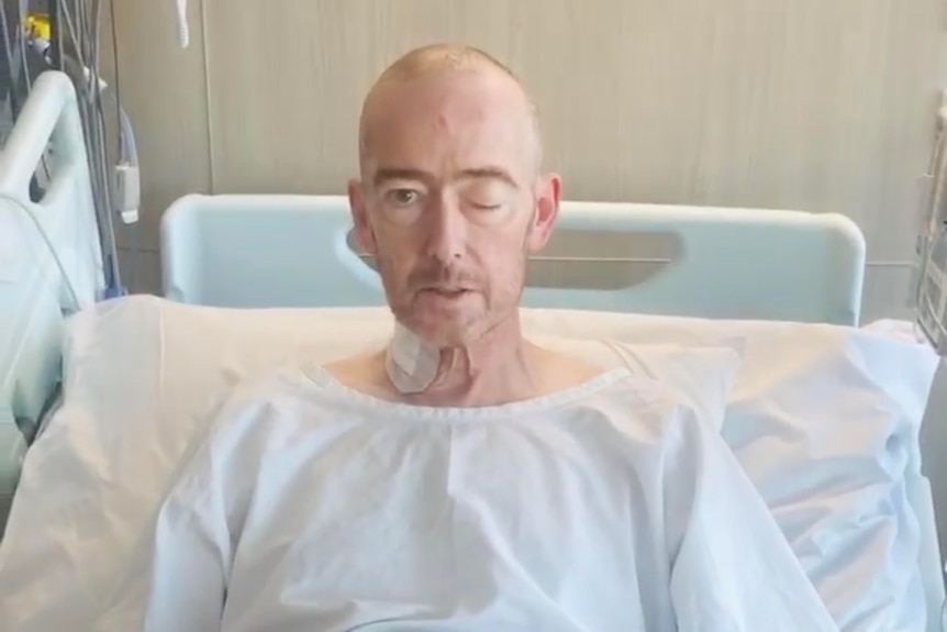 Man in hospital robe sits on bed with one eye closed