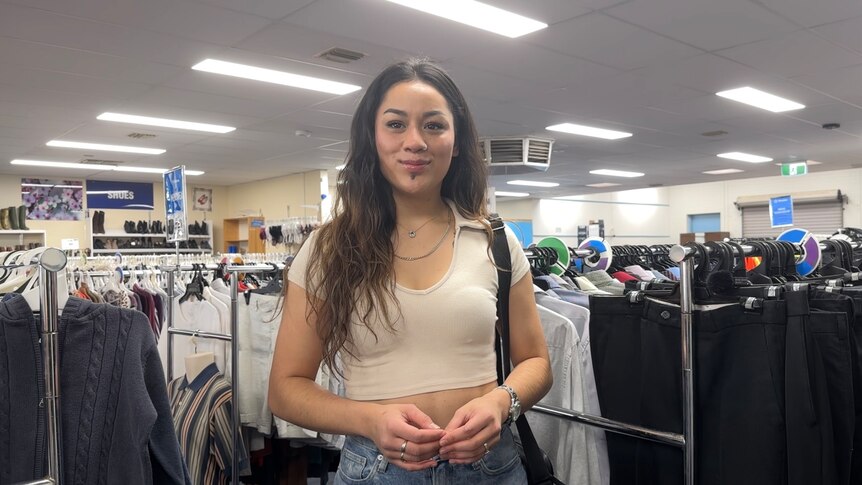A smiling young woman with dark hair stands in a second-hand clothing store.