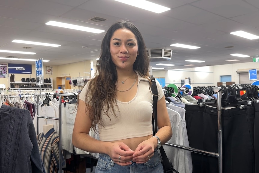 A smiling young woman with dark hair stands in a second-hand clothing store.