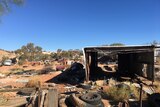 Rusted cars, old tyres, oil barrels and a derelict shed sit on a dusty red desert landscape