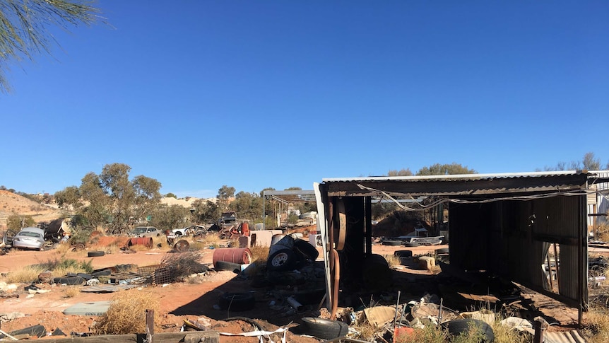 Rusted cars, old tyres, oil barrels and a derelict shed sit on a dusty red desert landscape