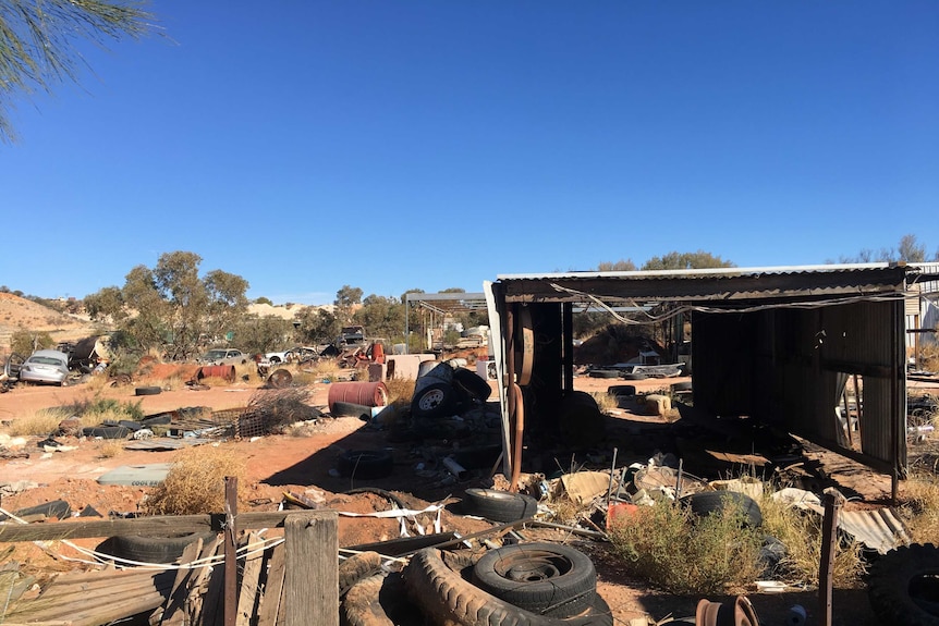 Rusted cars, old tyres, oil barrels and a derelict shed sit on a dusty red desert landscape.