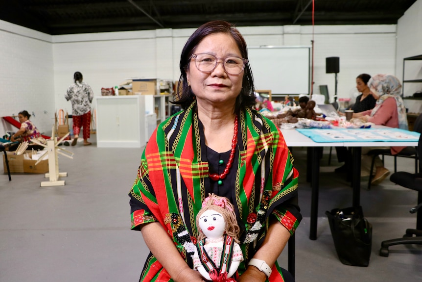 A mid-shot of a middle-aged Burmese woman sitting down indoors in a craft class posing for a photo while holding a doll.
