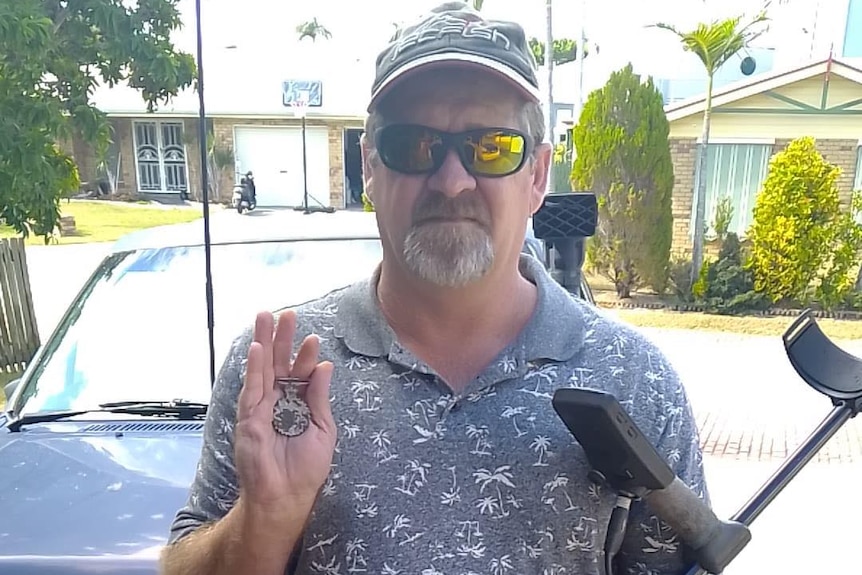 a man stands holding a medal in one hand and a metal detector in the other