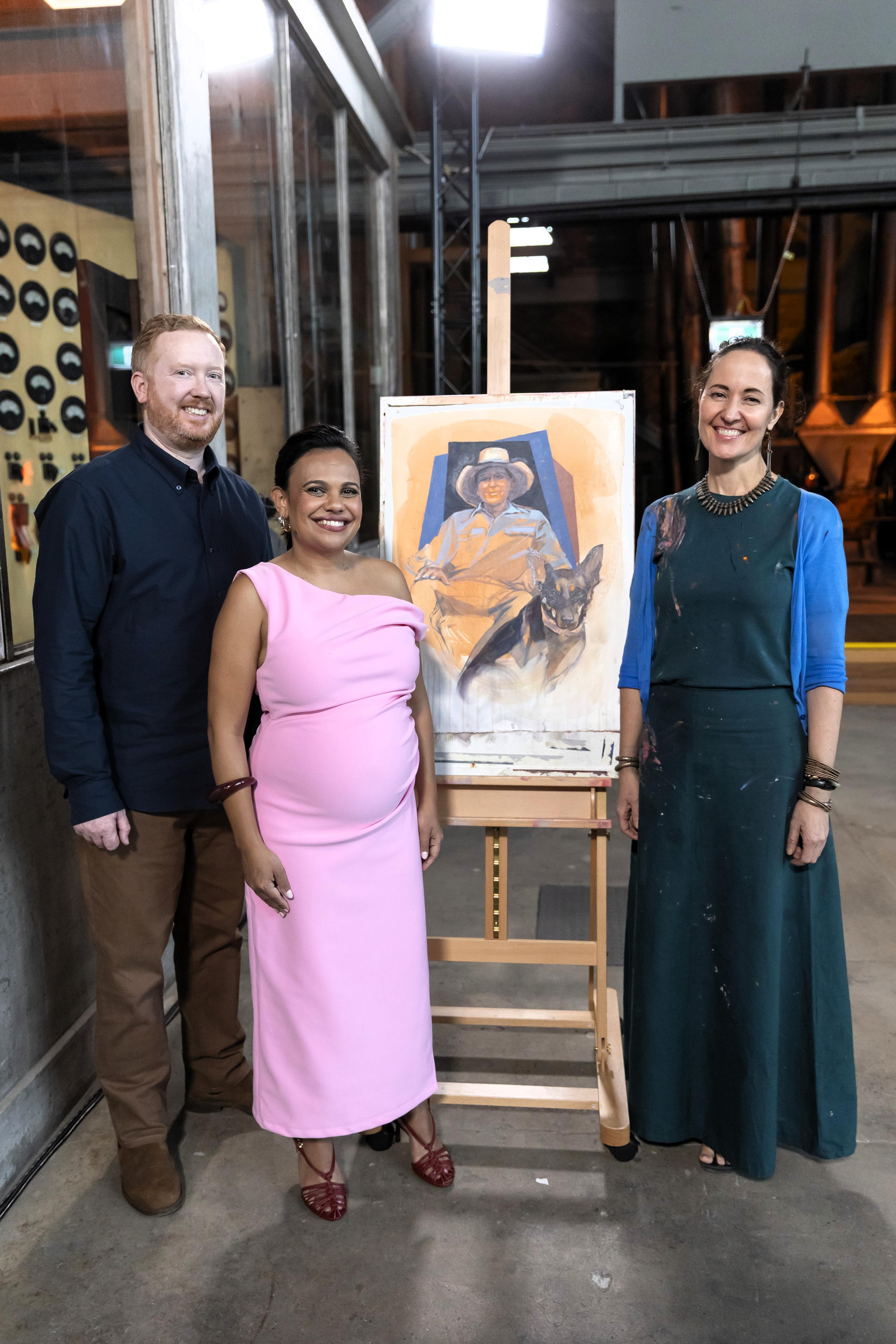 A man and two women stand in front of a portrait of a man and dog.