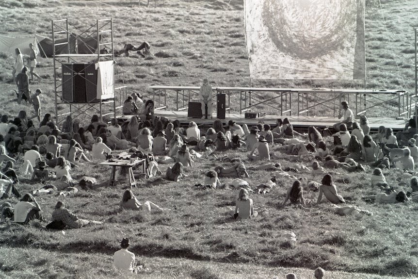 A black and white photo of people sitting around a small stage outside on the grass 