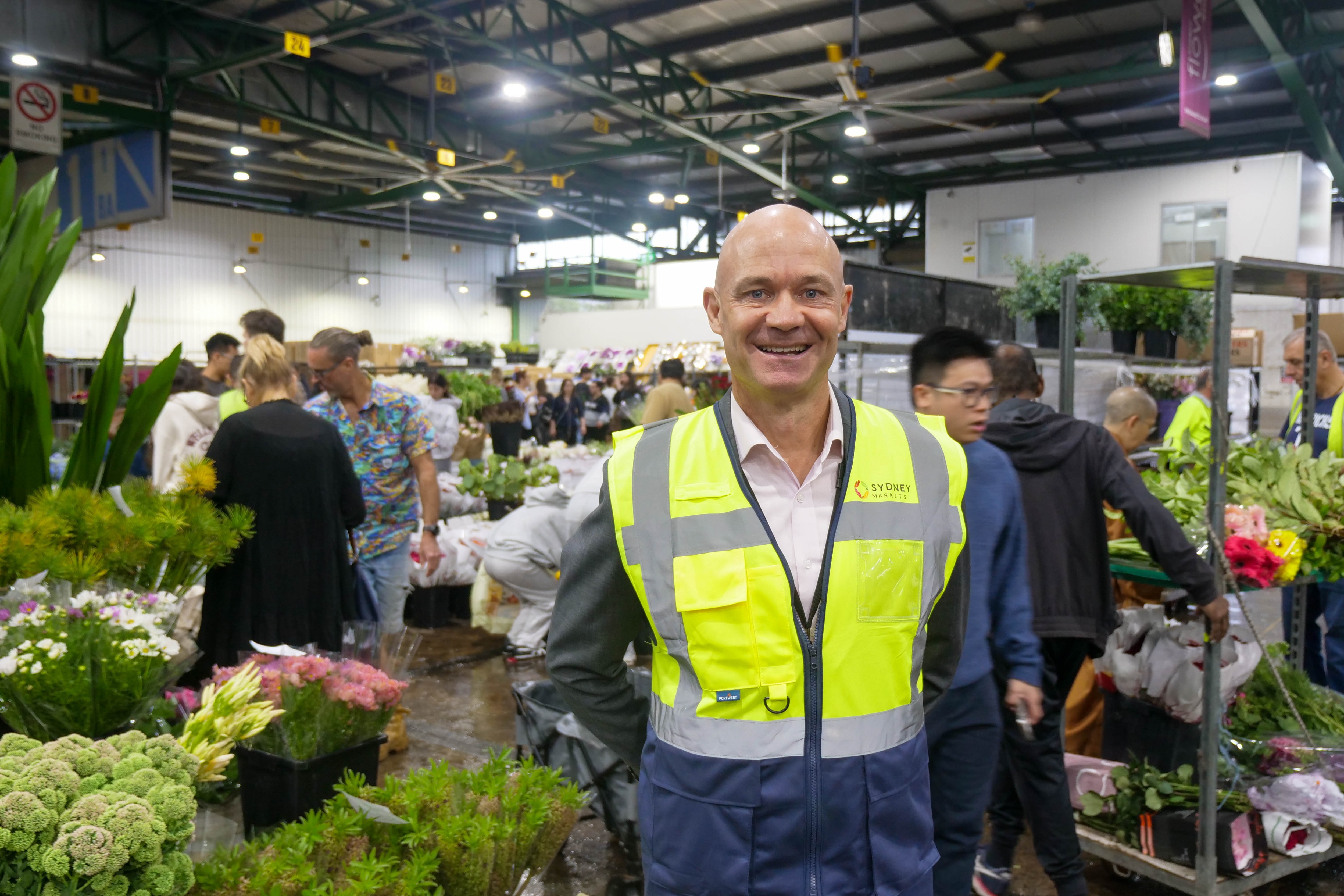 Un hombre calvo con un chaleco reflectante sonríe en un mercado concurrido