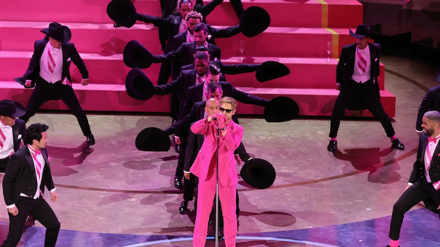 Ryan Gosling in a pink suit in front of men in suits on a stage. 